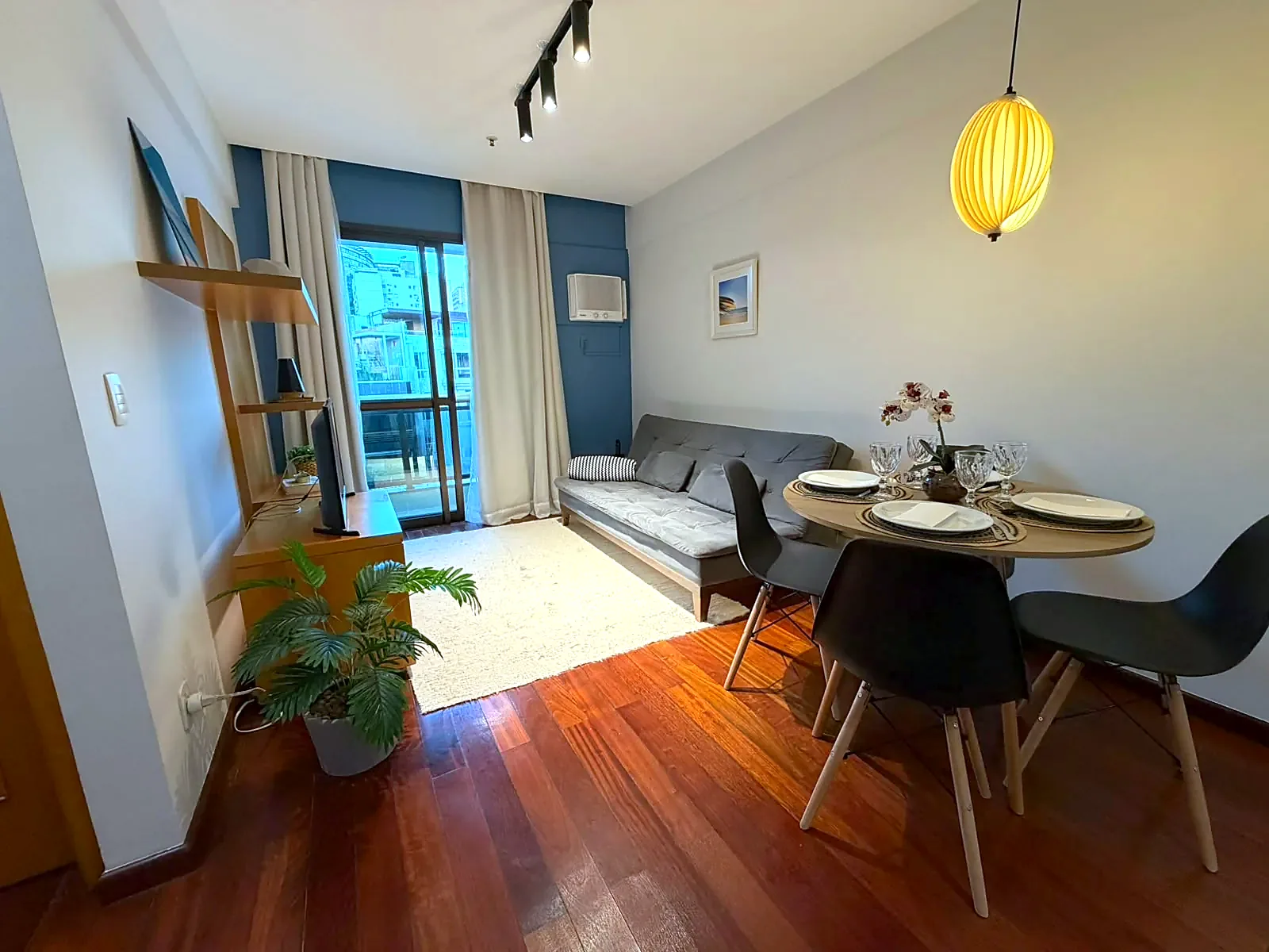 Sala de estar e jantar do Flat Maré Boa em Ipanema, com sofá, mesa posta para quatro, luminária de destaque e acesso à varanda com vista